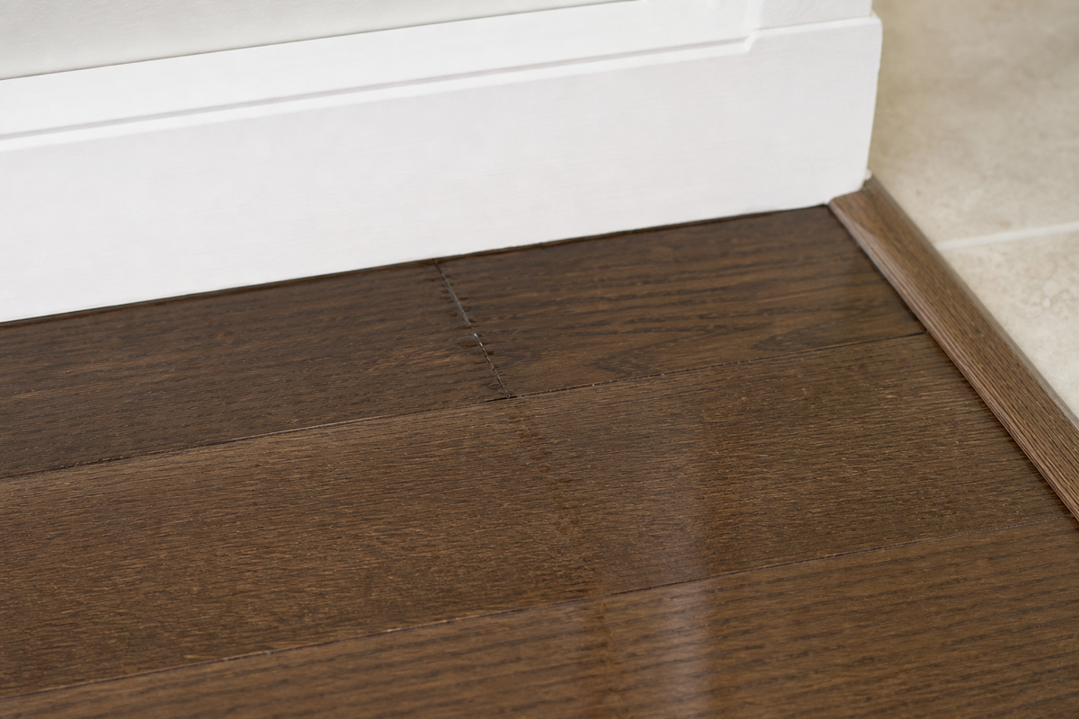A subtle damp path along a sealed hardwood plank seam beside a baseboard and transition edge.